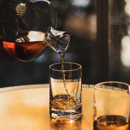 Close-up of a hand holding the Alba crystal whisky glass filled with amber spirit and a single ice sphere.